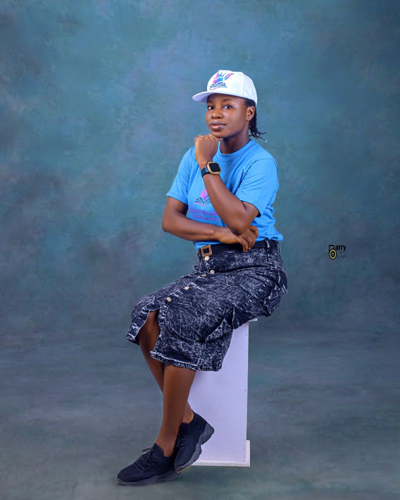 A young person sitting on a white pedestal, wearing a blue t-shirt, black denim shorts, and a white cap, with a thoughtful pose against a textured green background.