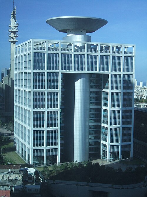 Modern architectural building with a distinctive circular top design and a central opening, surrounded by green landscaping.