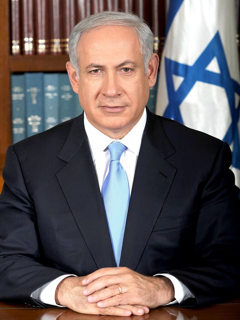 Portrait of a man in a suit with a blue tie, seated at a desk, hands folded, with bookshelves and an Israeli flag in the background.