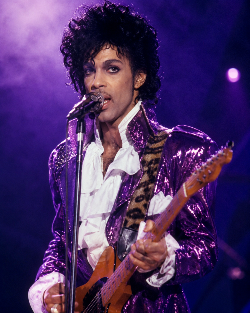 A male performer with curly hair, wearing a shiny purple jacket and a white ruffled shirt, singing into a microphone while holding an electric guitar on stage.