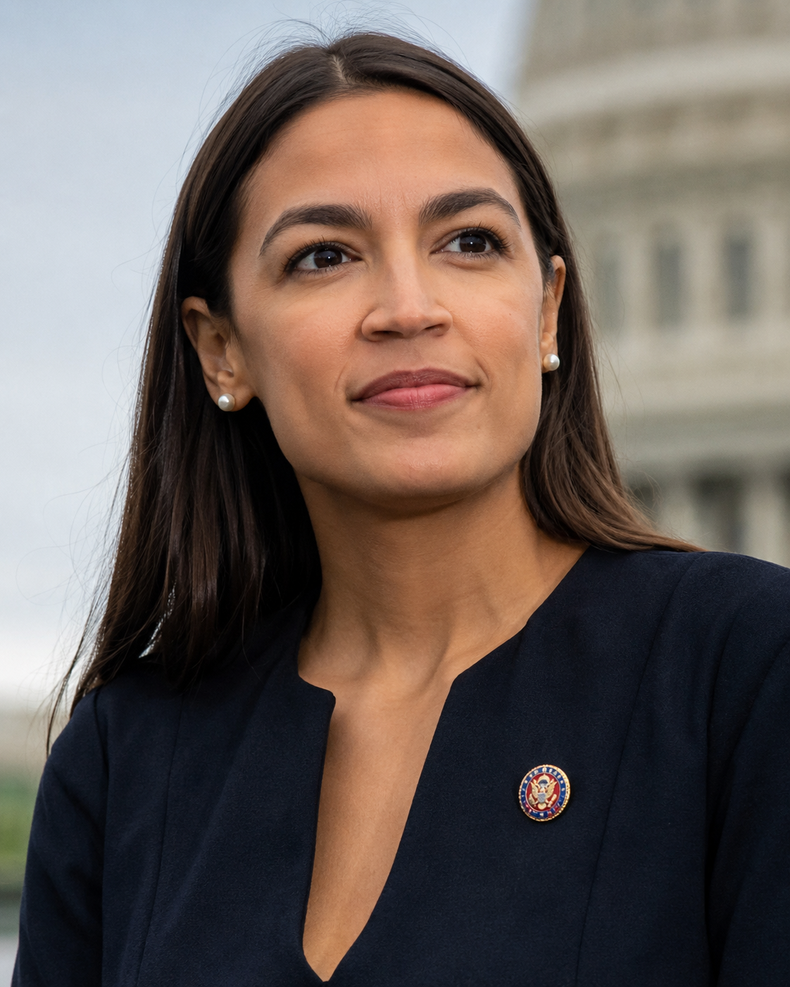 Close-up of a woman with long dark hair, wearing a dark blazer and a pin on her lapel, looking thoughtfully into the distance with a building in the background.