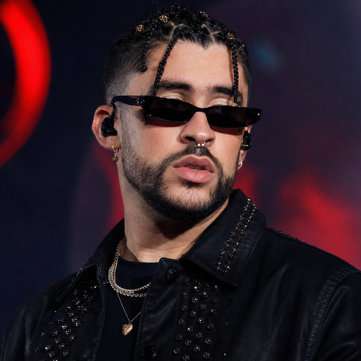 A close-up portrait of a man wearing sunglasses and a black studded jacket, with braided hair and facial piercings.