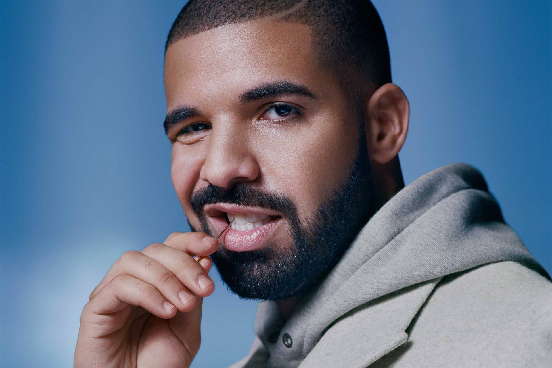 A close-up portrait of a man with a beard, looking at the camera while biting his finger, against a soft blue background.