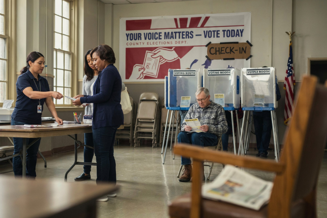 A busy polling place showing voters checking in and casting their votes. Two women are conversing with a poll worker at a table, while an older man reads a newspaper in the background near voting booths. A banner encourages voting, emphasizing its importance.
