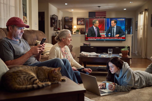 A family sitting in a living room, with a man using a smartphone, a woman engaging with a laptop, and an older woman watching a news broadcast on the television. A cat is resting on the couch.